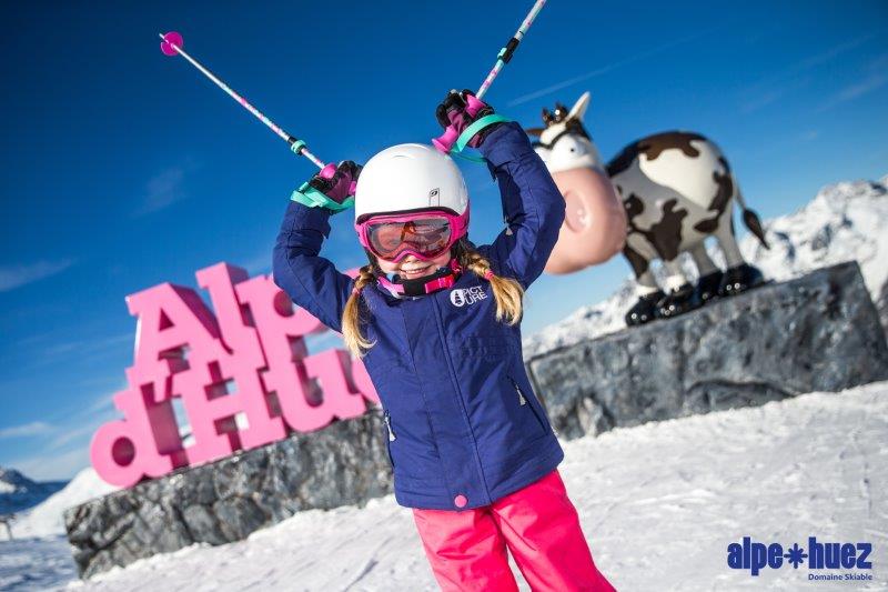 A junior skier stands in front of the Alpe D'Huez Ski Resort sign