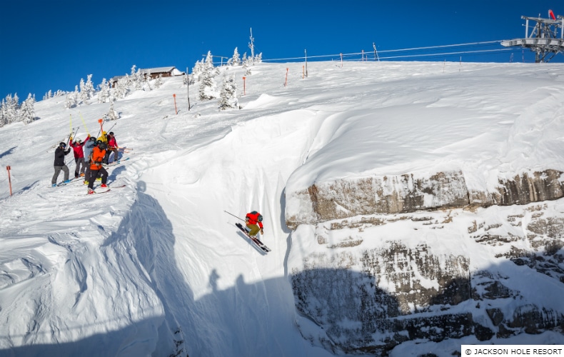 A skier in red drops into a steep slope at the top of a mountain