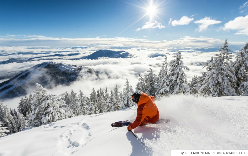 Skier in red and orange jacket skiing at Red Mountain, looking across the mountain range against a stunning blue sky